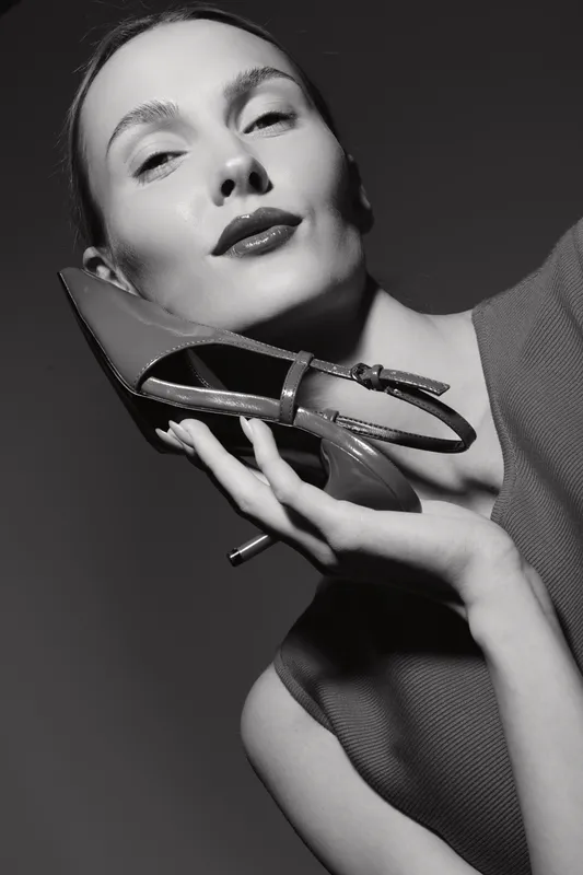 Portrait of a fair skinned Caucasian woman holding a high heel shoe near her face with a confident expression. Black and white studio photo emphasizing fashion and elegance.