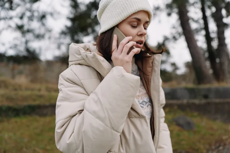 Woman in a light beige winter coat talks on her phone outdoors in a park, bundled in a knit hat, autumnal grass and trees in soft focus