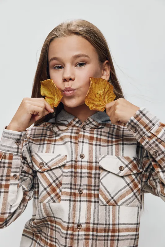 Girl holding two dried leaves near face making playful expression in casual plaid shirt isolated on white background. Child with autumn mood smiling and having fun with foliage indoors.