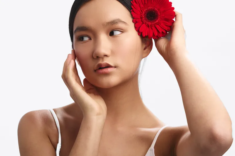 asian woman gracefully poses with a red flower tucked behind her ear, soft studio lighting highlights smooth skin and a serene expression, minimal makeup, white garment, creating an elegant and