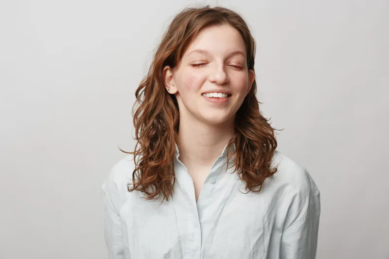Young woman with curly hair smiles with closed eyes wearing white shirt on light background. Concept of natural beauty, happiness, casual style, and relaxed mood in studio portrait.