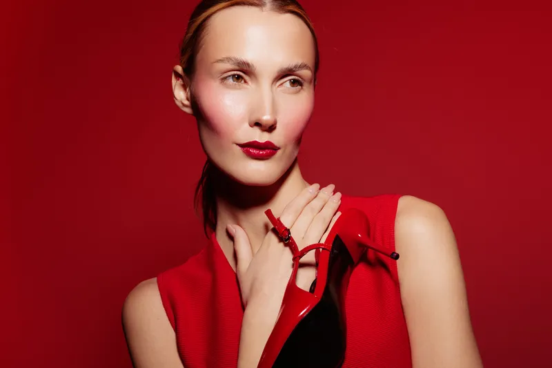 Elegant woman in red dress holds red high heel shoe near her neck against red background. Fashion concept with stylish makeup and confident pose in studio lighting.