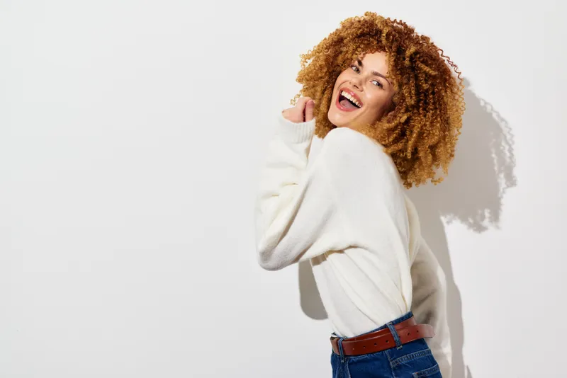 Радостная молодая женщина с красивыми вьющимися волосами ярко улыбается на фоне однотонного белого фона