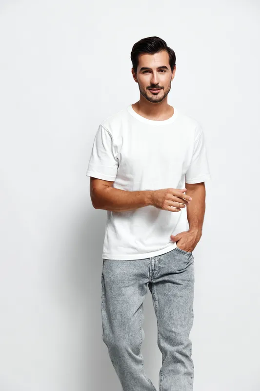 Full body smiling young man wearing white t-shirt and grey pants, looking at the camera while holding a cigarette in hand, isolated on plain solid background. Casual lifestyle concept.