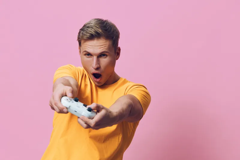 excited young caucasian man wearing yellow shirt holding white game controller playing video game isolated on pink background digital detox concept