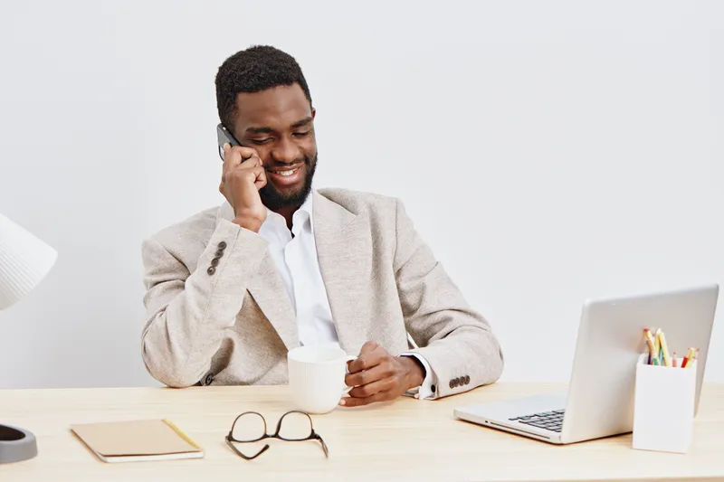 Professional smiling man talking on mobile phone while sitting at desk with laptop, notebook, and stationery in bright office. Business communication, remote work, technology, workplace lifestyle conc