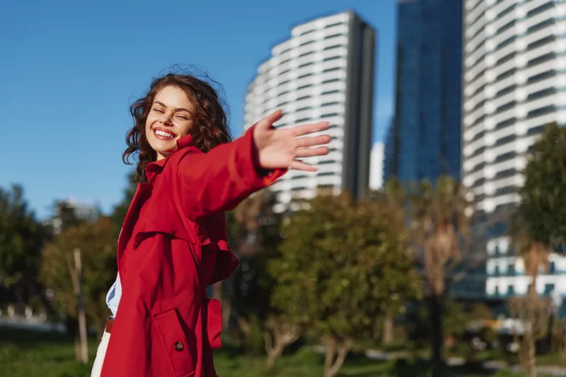 Joyful woman outdoors, red coat, arms outstretched, sunlit smile, urban park scene with tall buildings, blue sky, natural colors, carefree moment, candid portrait
