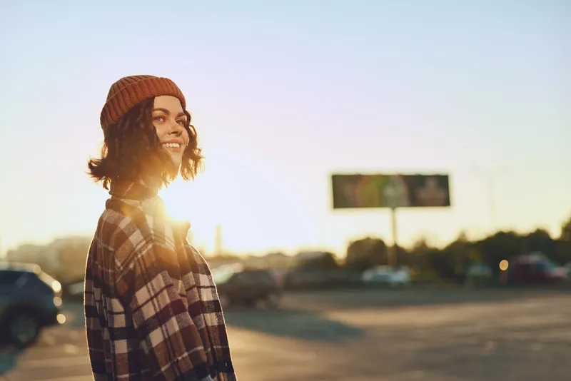Woman smiling in beanie and plaid shirt at golden hour, sunlight backlight in an urban parking scene. Candid lifestyle portrait with authenticity, golden hour glow and mindful living.