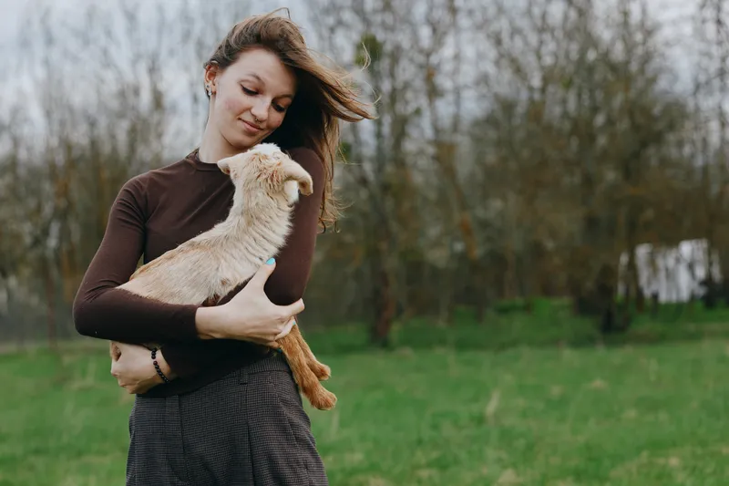 Caring woman holding a goat kid in a grassy park, outdoor moment, candid lifestyle scene, gentle interaction, warm daylight, sense of companionship and rural charm.