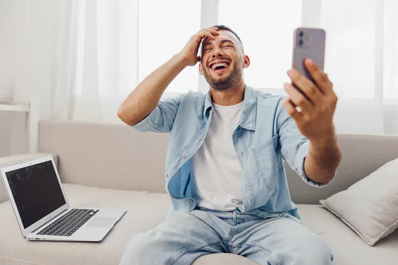 Happy man enjoying a digital detox while laughing during a phone call at home, embodying emotional minimalism and soft masculinity in a cozy setting.