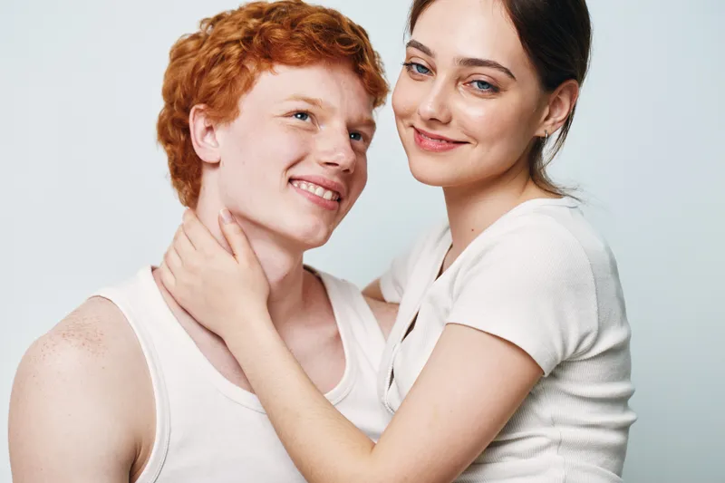 Couple, young man and woman smiling together with close embrace, casual clothing and positive mood in studio with light background and natural expression of happiness.
