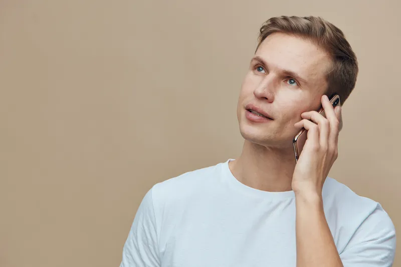 Close-up of a smiling young Caucasian man with short light brown hair wearing a white t-shirt looking away while talking on a mobile phone, isolated on plain beige background. People lifestyle concept