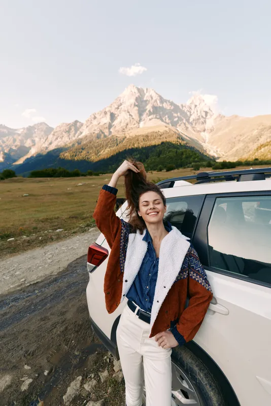 woman car mountains travel smile road nature adventure portrait of a happy female by an SUV, wearing casual jacket and denim, standing on a scenic roadside enjoying outdoor freedom.