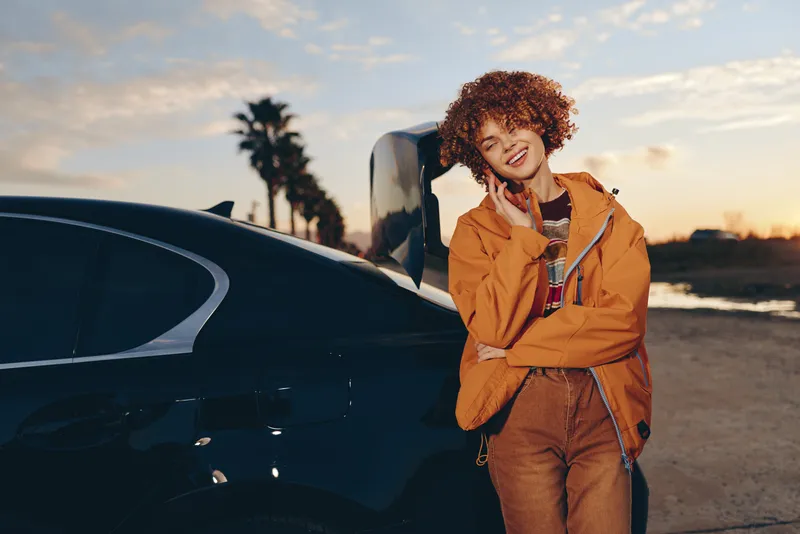 Woman lifestyle outdoors near car at sunset, smiling with curly hair, wearing orange jacket and casual travel outfit, relaxed stance on roadside, warm sunlight, soft breeze