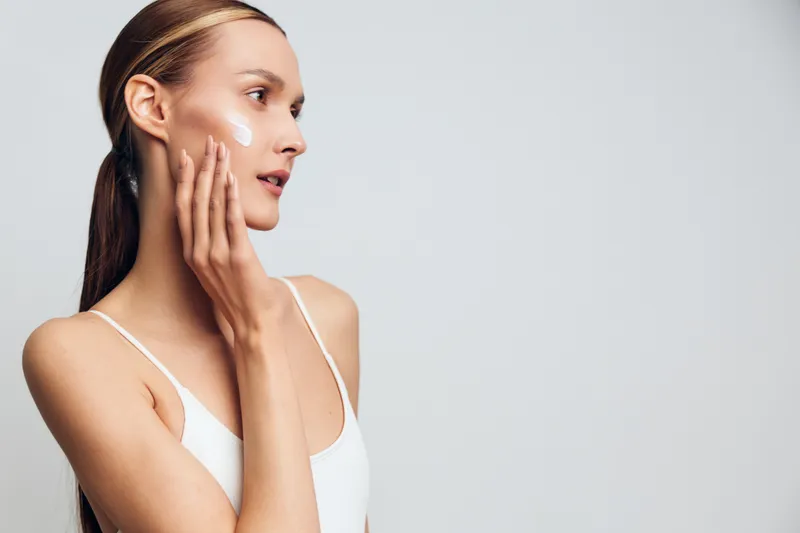 Young woman face cream skincare beauty self care moisturizing hydrated skin, hand on cheek, side profile, clean studio portrait, light gray background, isolated