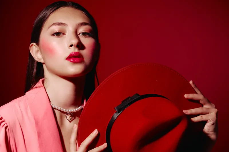 Woman in pink jacket with pearl necklace poses with red hat and bold glossy lips. Her makeup has dramatic rosy blush. Fashion shot with a luxurious and confident beauty concept.