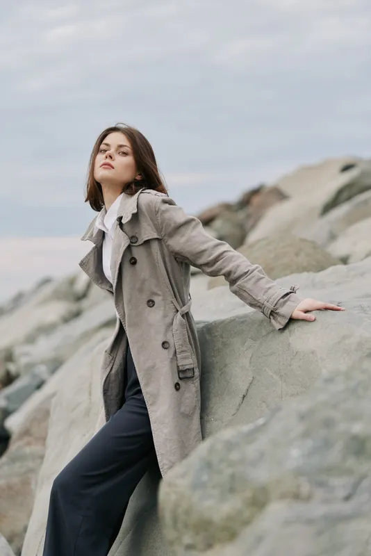 Young woman in long coat poses on rocky shore under cloudy sky. Fashion model leans on stone with confident look. Concept for autumn style and natural landscape.
