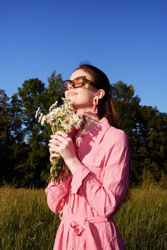 Young woman in a pink dress holding flowers, smiling under a blue sky, with a vibrant natural background, evoking feelings of joy and springtime
