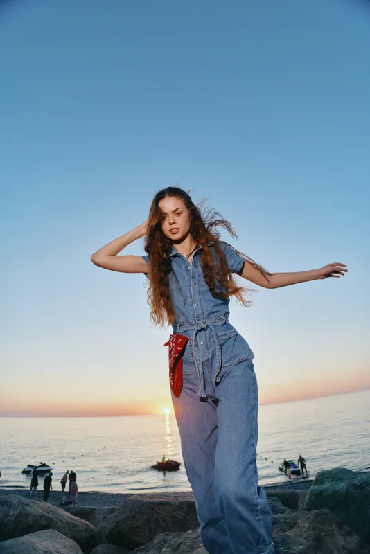 young woman in denim jumpsuit dancing on rocky beach at sunset with ocean background and warm natural light