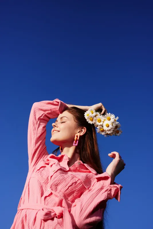 Stylish young woman wearing a pink dress and flower crown against a blue sky, embodying joy and summer vibes with a cheerful expression