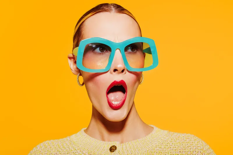 Surprised woman with bright red lipstick and large blue sunglasses looks shocked against vibrant yellow background. Fashion concept with bold colors and expressive facial expression.