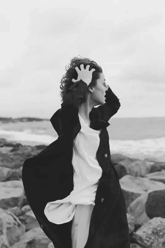Windy ocean scene with a coat fluttering, a woman on rocky shore expressing freedom and motion against a gray seascape