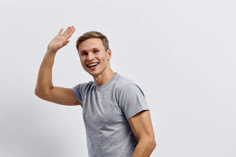 Smiling man young adult casual grey t-shirt waving hand friendly greeting isolated on plain white background studio portrait people happiness concept
