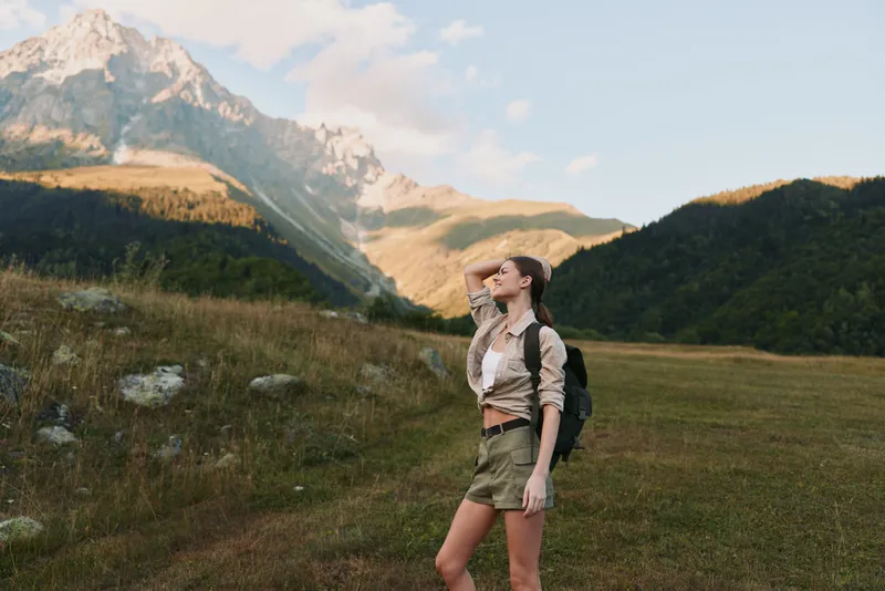Woman outdoors hiking with backpack, admiring mountains landscape under sunshine, relaxed posture, nature mood, travel vibe, cheerful expression, scenic terrain
