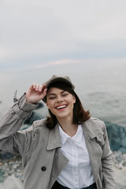 Young woman wearing vintage coat and cap smiles joyfully while standing on rocky beach. Overcast sky and calm sea create a relaxed natural background. Concept of happiness and fashion.