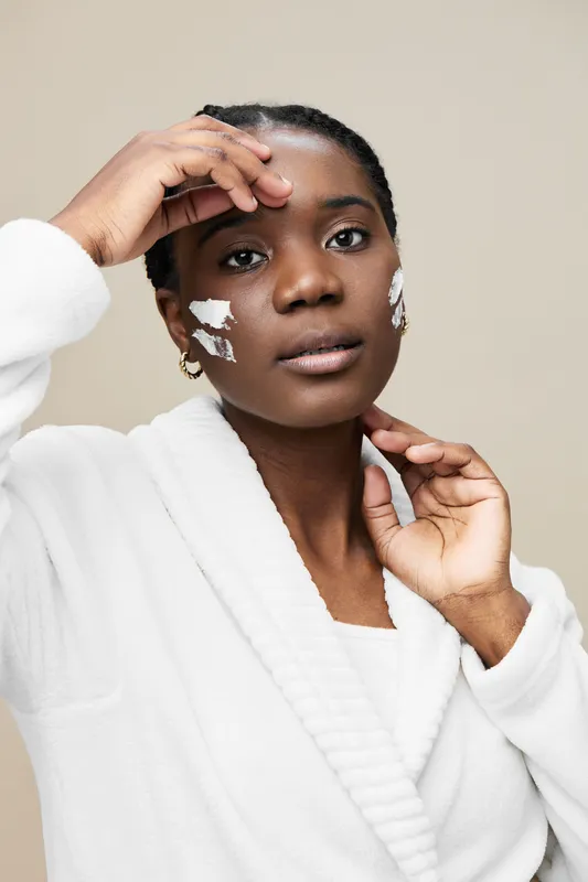 Young Black woman applying skincare cream, showcasing a natural glowing complexion, wearing a white robe against a soft beige background
