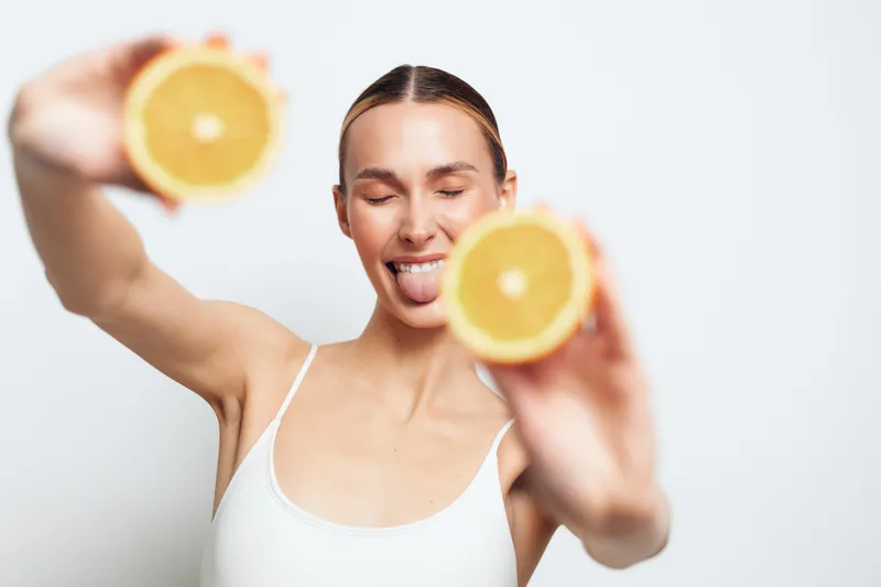 A smiling young woman with tied back hair holds two orange halves in front of her face, sticking out her tongue playfully. The background is plain white, emphasizing freshness and fun.