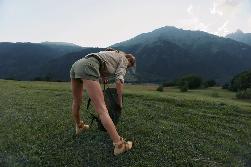 A woman hiking outdoors bends to adjust a backpack in a green meadow, with distant mountains and clear sky, depicting hiking, exploration, and nature lifestyle moments.