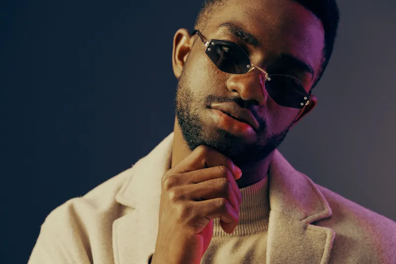 Stylish young African man in sunglasses and elegant outerwear posing thoughtfully against a colorful backdrop, showcasing confidence and modern fashion trends