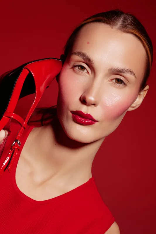 Close-up of a woman with red lipstick and blush holding a red high heel shoe near her face. Red background and dress create a bold fashion and beauty concept with strong color contrast.