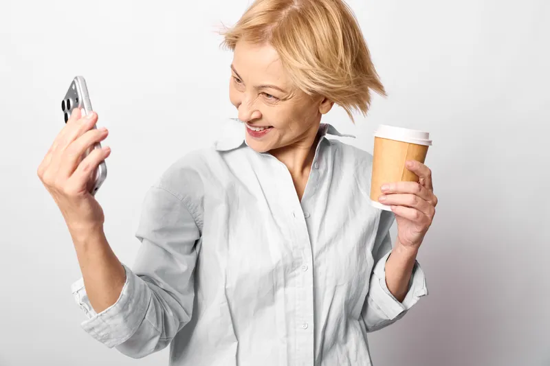 Senior woman indoors holding smartphone and coffee cup, smiling softly, bright white background, casual light shirt, relaxed cheerful moment at home