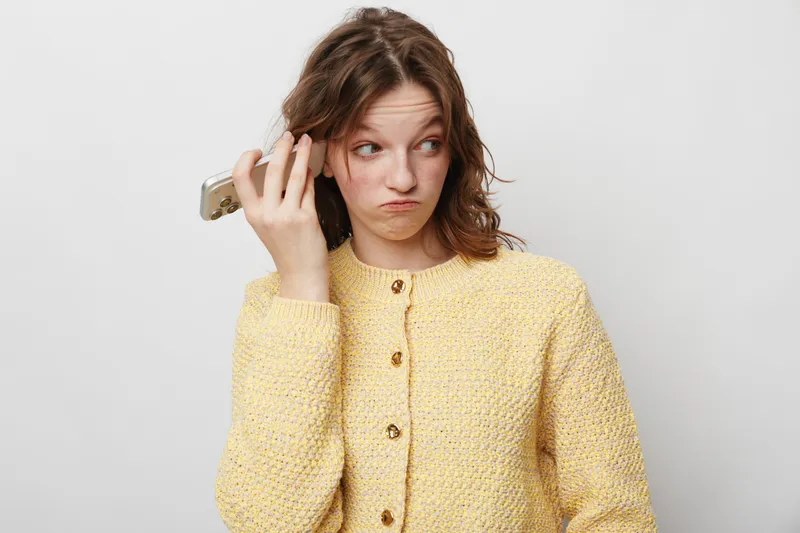 Young woman in yellow knitted sweater holds smartphone near ear with confused expression. Isolated on white background. Concept of misunderstanding, communication, and technology frustration.