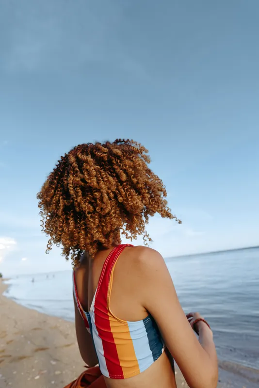 Woman curly hair beach ocean swimsuit sunlight summer relaxation, viewed from behind at shoreline, red striped suit, sunlit calm waves, relaxed posture, natural blues soft breeze today