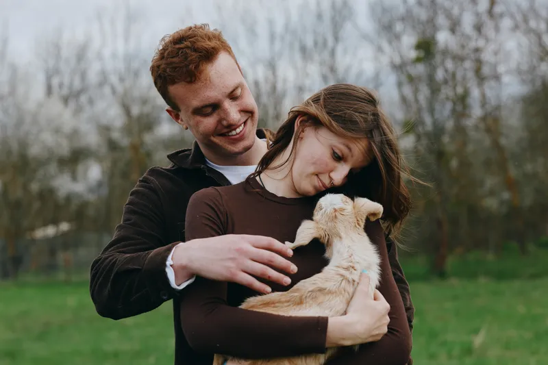Couple in warm casual clothes smiling as they cradle a gentle goat kid in an open field, showcasing a cozy lifestyle moment full of affection and outdoor happiness.