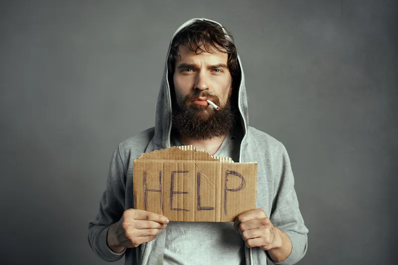 Бородатый мужчина в капюшоне с сигаретой держит картонку с надписью help