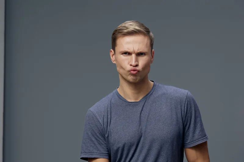 Young man making silly face, pouting lips, serious expression, casual grey t-shirt, short hair, light skin, studio shot against plain grey background. People emotion concept