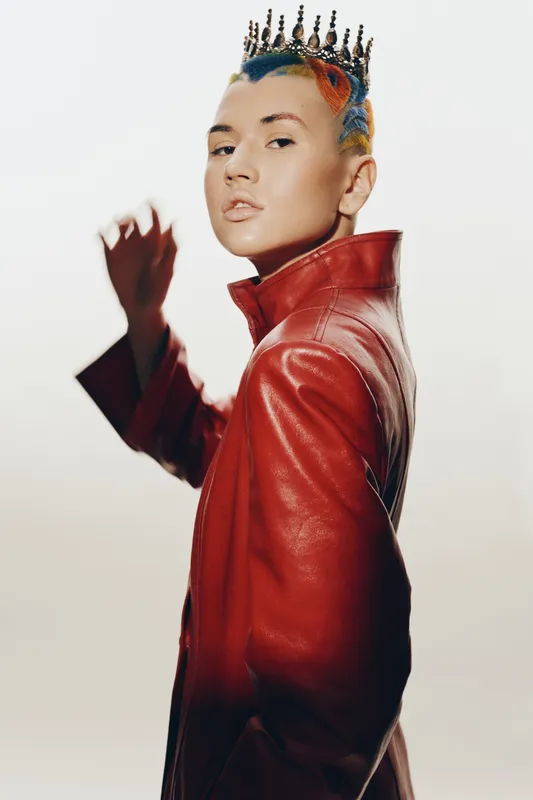 Young woman with colorful hair wearing a red leather jacket and a crown accessory, posing confidently against a soft, neutral background in a modern fashion style.