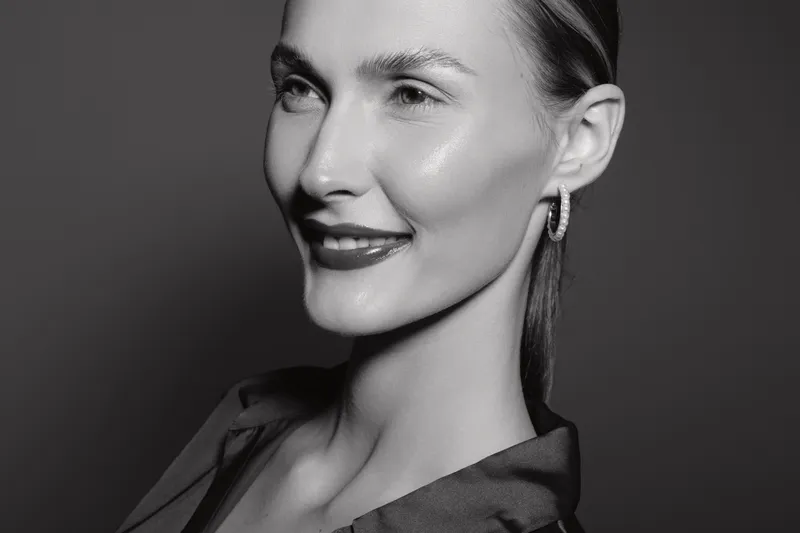 Portrait of a smiling Caucasian woman with fair skin and slicked-back hair wearing hoop earrings and a dark blouse. Studio lighting highlights her clear complexion and confident expression.