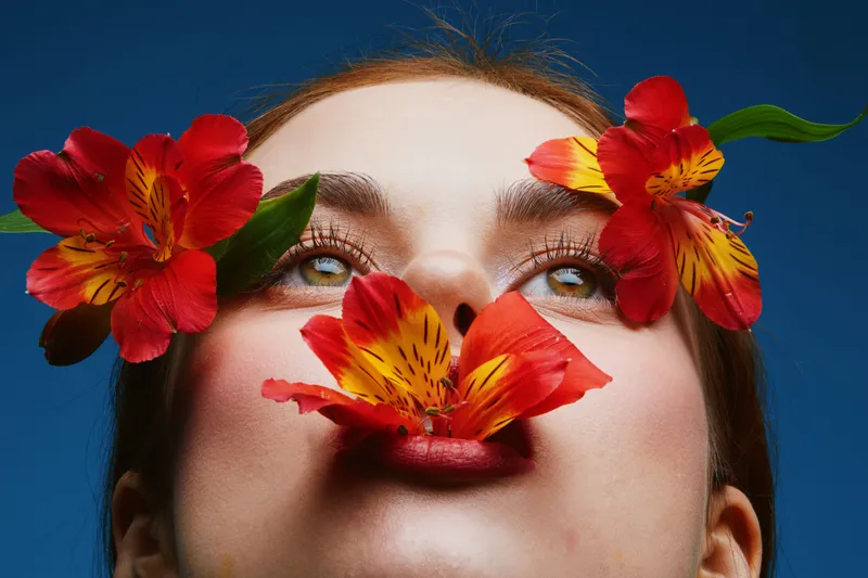 Close up of a woman with vibrant flowers adorning her face, embodying beauty and nature s harmony, against a bold blue background, perfect for wellness themes