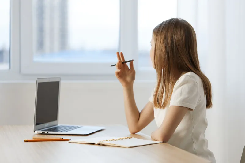 Thoughtful young woman working from home, sitting at a desk with a laptop, a notebook, and a pencil, contemplating ideas by the window in a bright, minimalistic environment