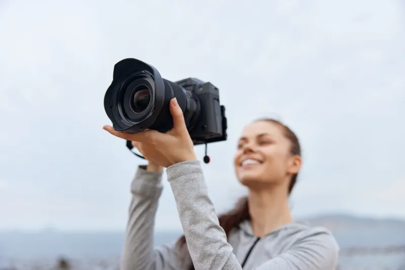 Smiling woman holding a professional camera outdoors, showcasing passion for photography with a cloudy backdrop, ideal for creative and lifestyle themes