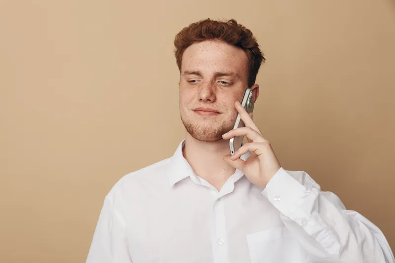 Young man with red hair wearing white shirt talks on smartphone, looking down with slight smile. Neutral beige background. Concept of communication, technology, and casual lifestyle.