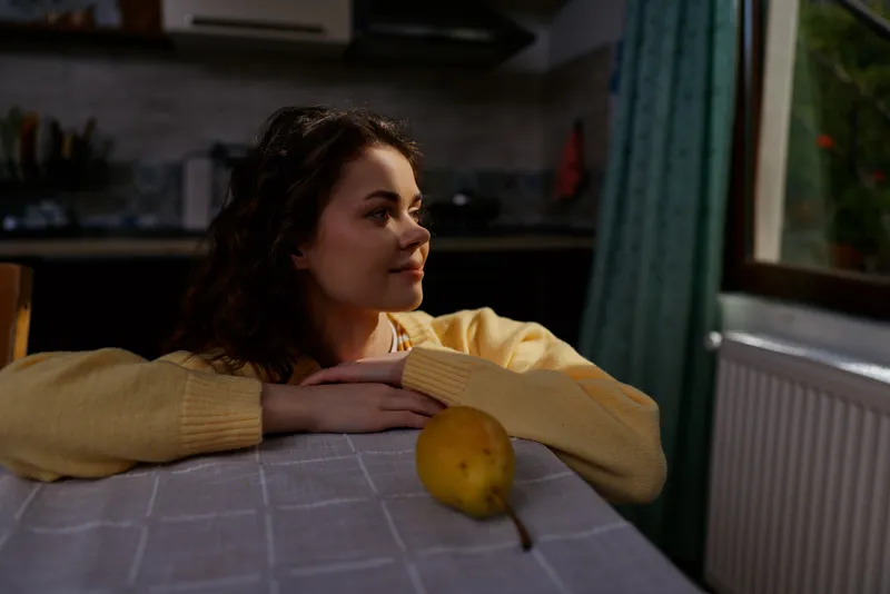 Cozy indoor moment with a woman resting on a kitchen table, warm sweater, soft daylight, calm expression, apple on the table, relaxed lifestyle at home, intimate everyday scene