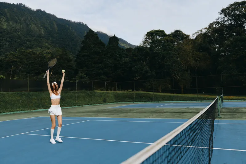 Young female tennis player celebrating victory on outdoor court, wearing sporty attire, surrounded by green mountains and blue sky, conveying excitement and achievement