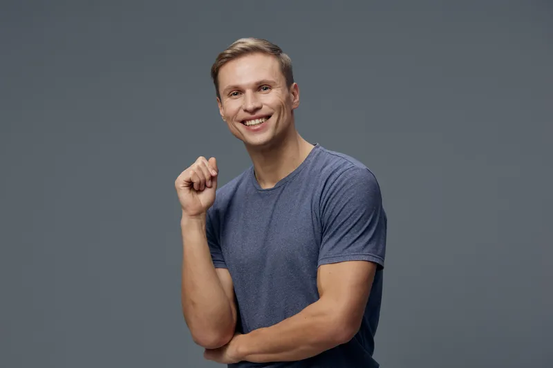 Cheerful young man with short light hair smiling confidently while wearing a casual blue t-shirt, posing with a fist gesture on a plain gray background. Studio portrait of happy male for lifestyle and
