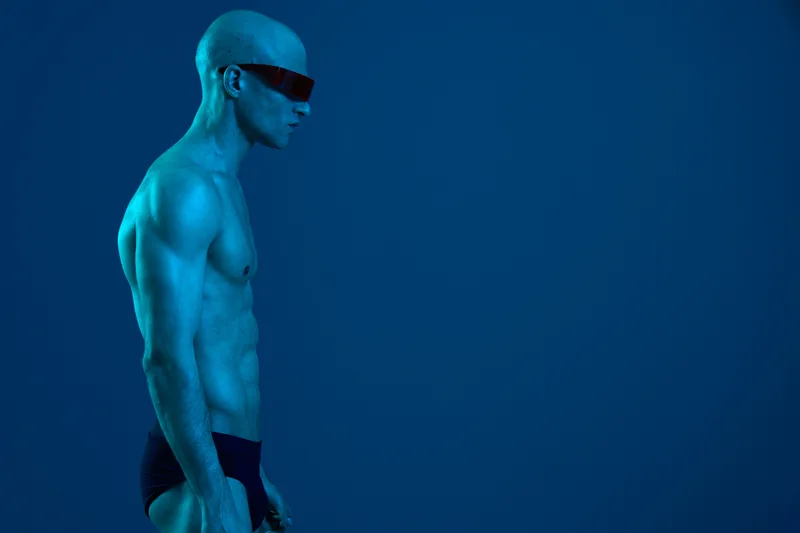 Athletic man posing in a blue light setting, showcasing his muscular physique and wearing sunglasses for an artistic black and white aesthetic.